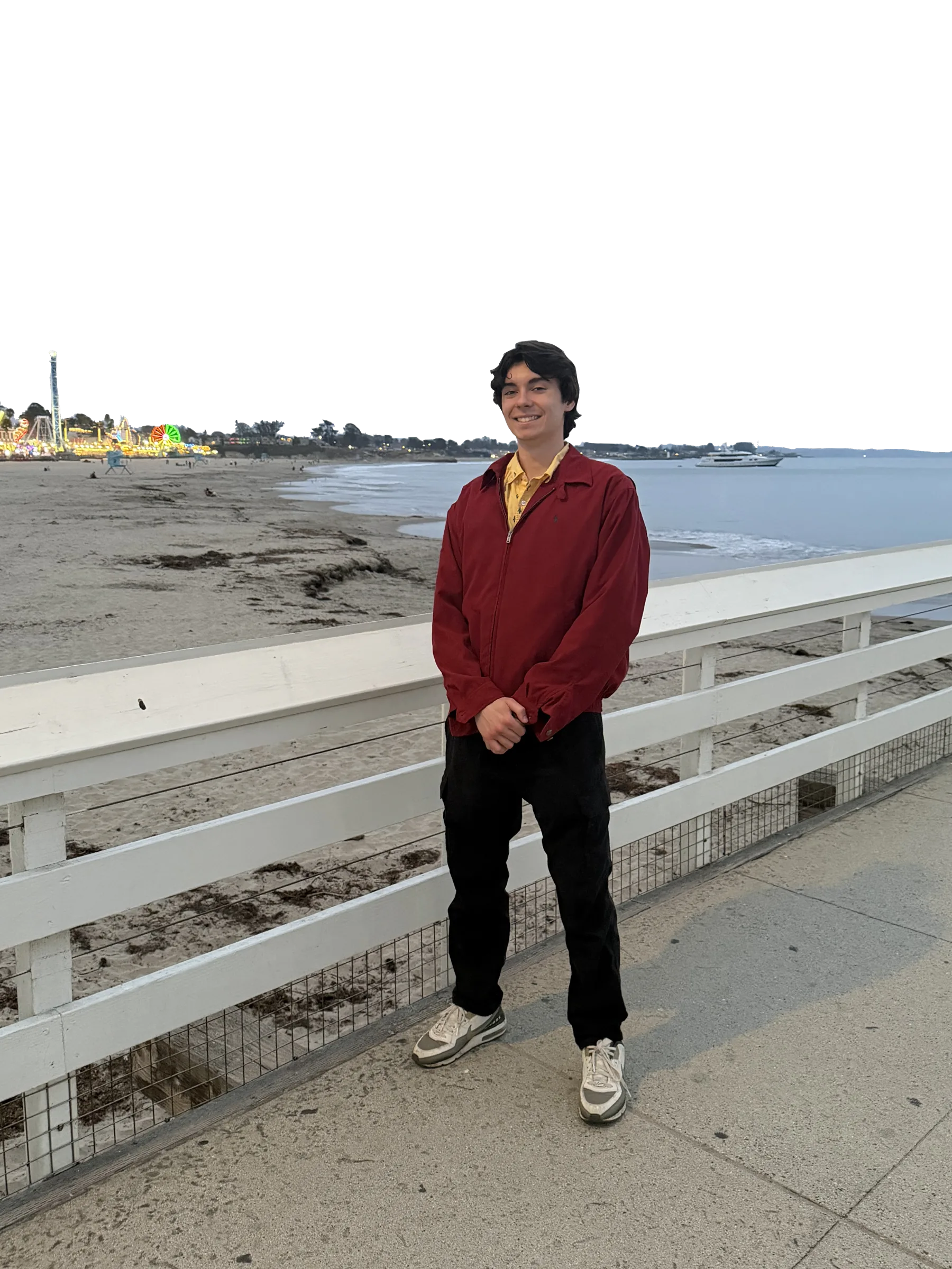 James smiling on a pier, wearing a red jacket, with the beach behind him